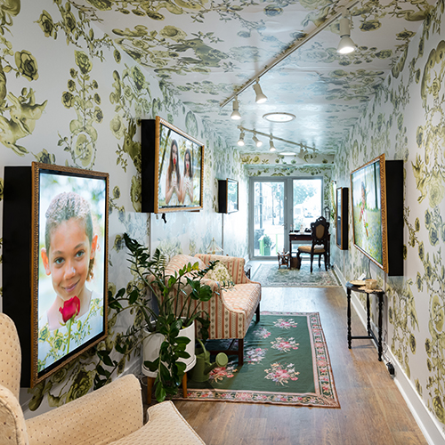 An art exhibit designed to look like a living room, with sofa, armchair, and video art hung on wallpapered walls.