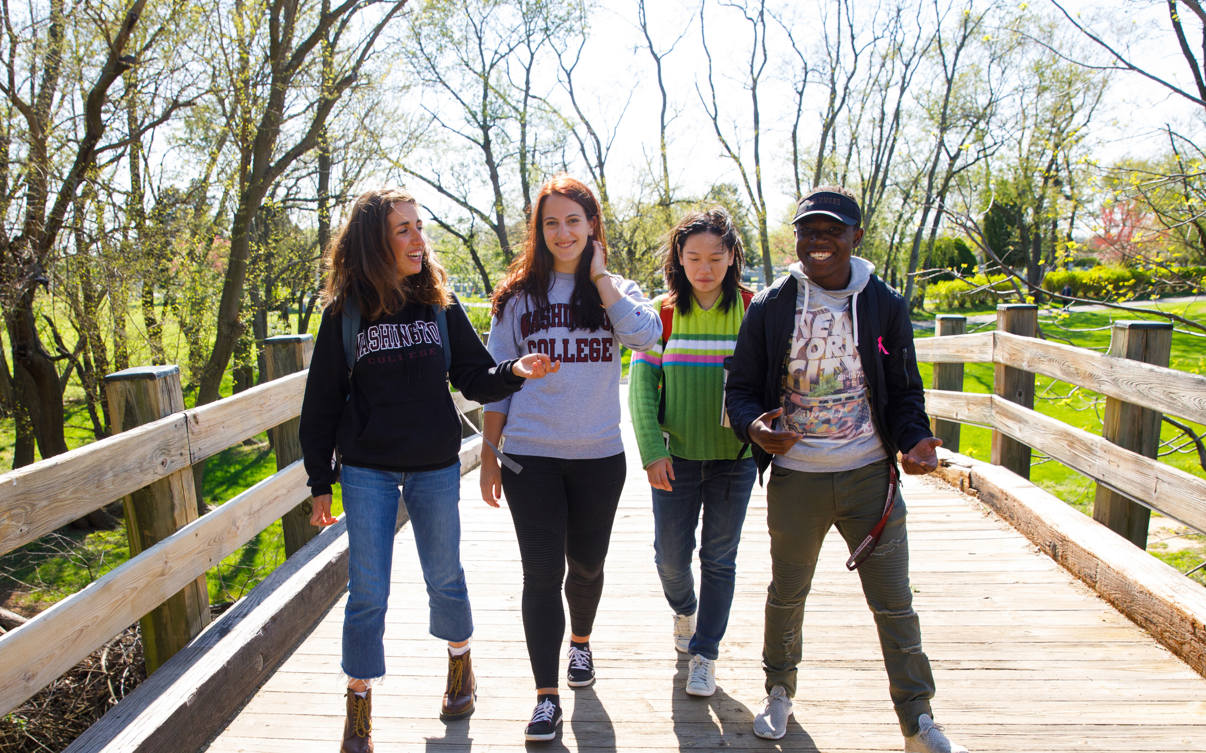 four college students; a black male, 2 white females, and an asian female, smiling and walking on a wooden bridge with green grass and trees in the background