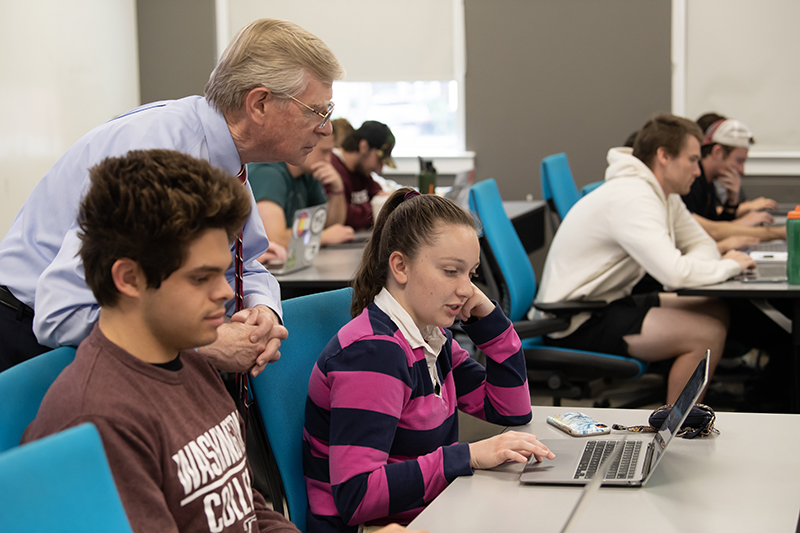 Professor Joe Bauer looks at the screen showing business simulation choices by Alex May '23, with Grayson Wright '25 alongside.