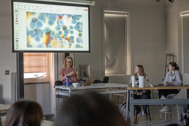 artist gestures while talking about her art, displayed in a powerpoint above her, to a room of students