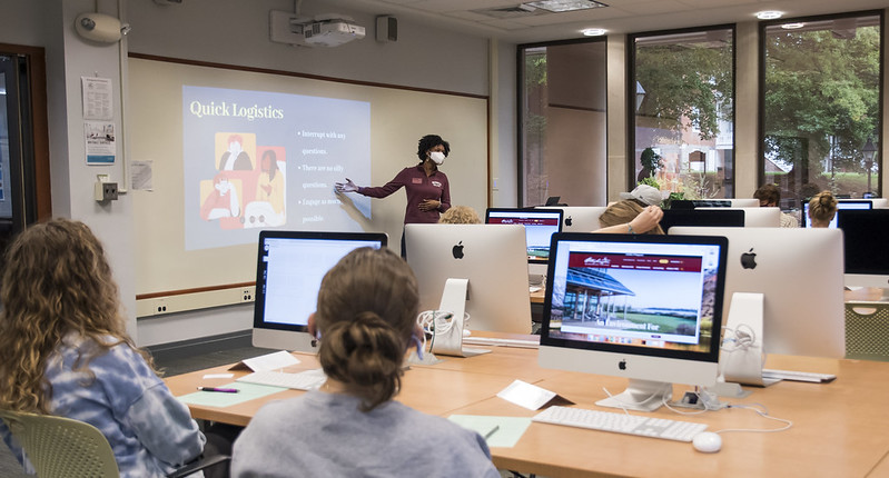 Students at computers look to an instructor at the front of the room reviewing a slide titled 