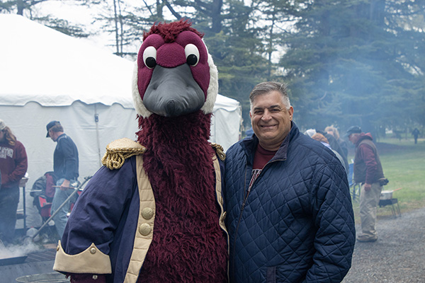 An alumnus poses with Gus the Goose in front of a cookout and party tent