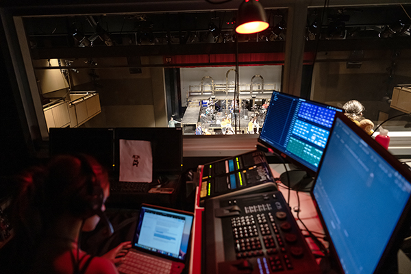 A student in the control booth at Decker Theatre with a performance just visible beyond her
