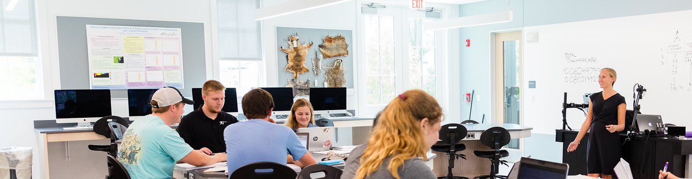 student seated at group tables listening to a faculty member lecture