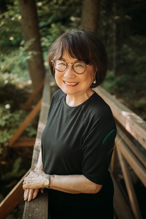 Kay Enokido smiles; the background is a copse of beautiful green trees