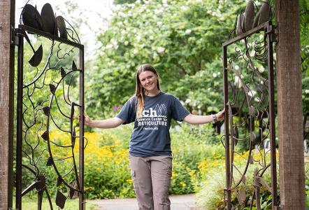 Shannon Roche '25 opens the gates at Delaware Center for Horticulture while facing the camera in a DCH tee-shirt with the garden behind her