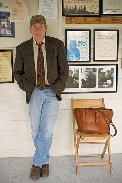 Robert Day in jeans, a tie, sportcoat and baseball cap, leans against a wall covered in framed literary memorabilia.