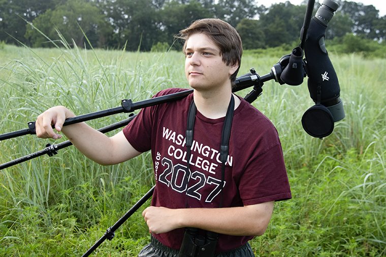 Justin Looper ’26 with a spotting scope over his shoulder standing in a field at the River and Field Campus.