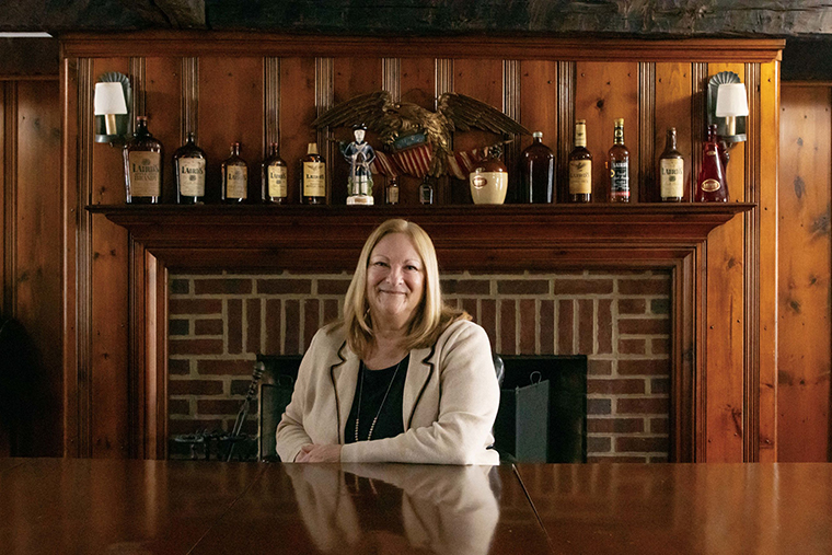 Lisa Laird Dunn ’83 sits at a table in front of a fireplace where bottles of her family’s liquors line the mantle