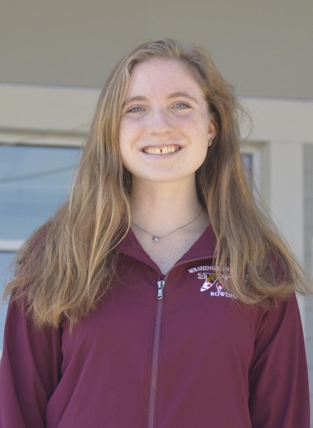 Sophia, wearing a maroon rowing coat and smiling with the boathouse behind her. 