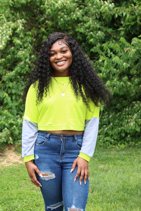 Ama, smiling and wearing a bright green shirt and blue jeans with trees behind her. 
