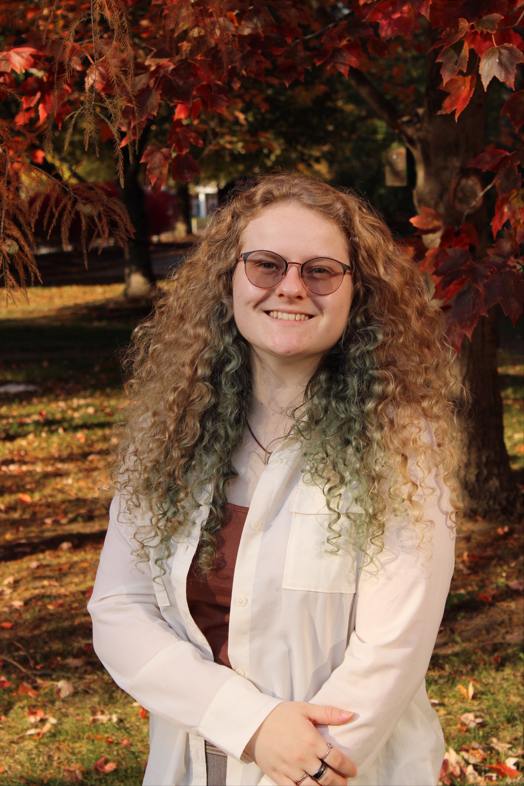 Arianne, smiling and wearing a white shirt with fall foliage behind her. 