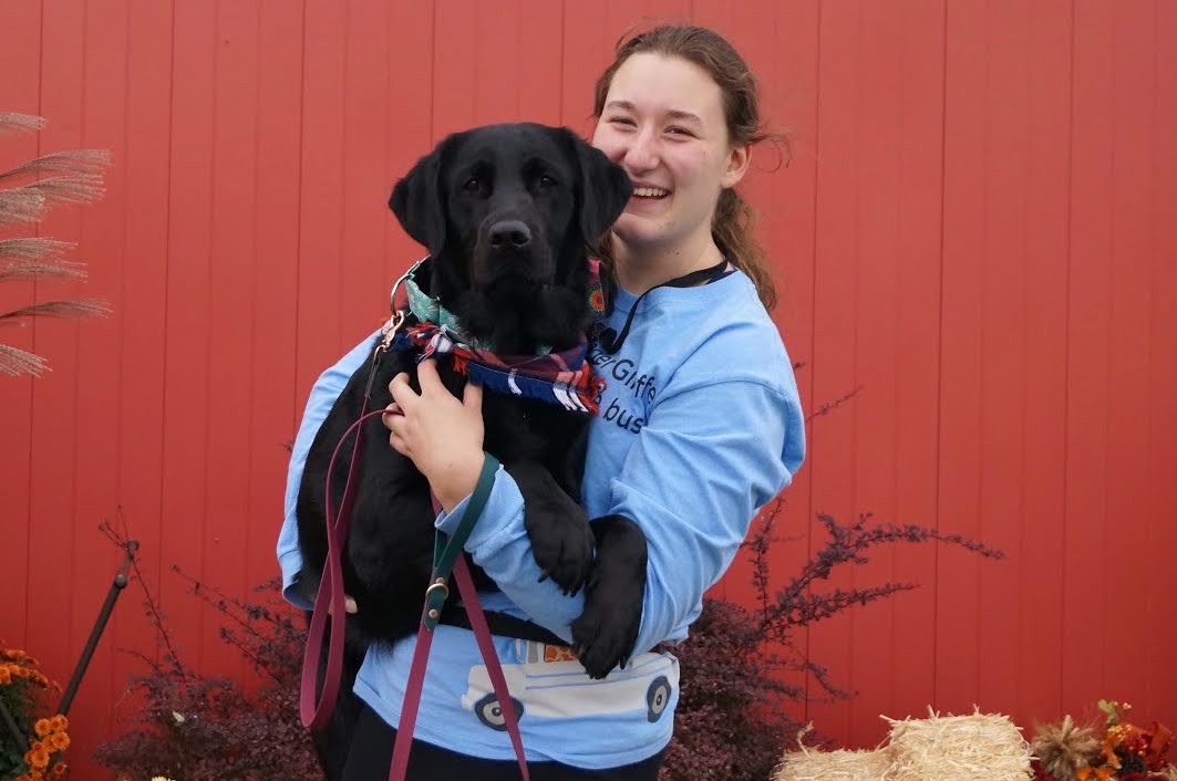 Grace, smiling and wearing a blue sweatshirt, holding a black lab dog with a red blue bandana in front of a red barn.