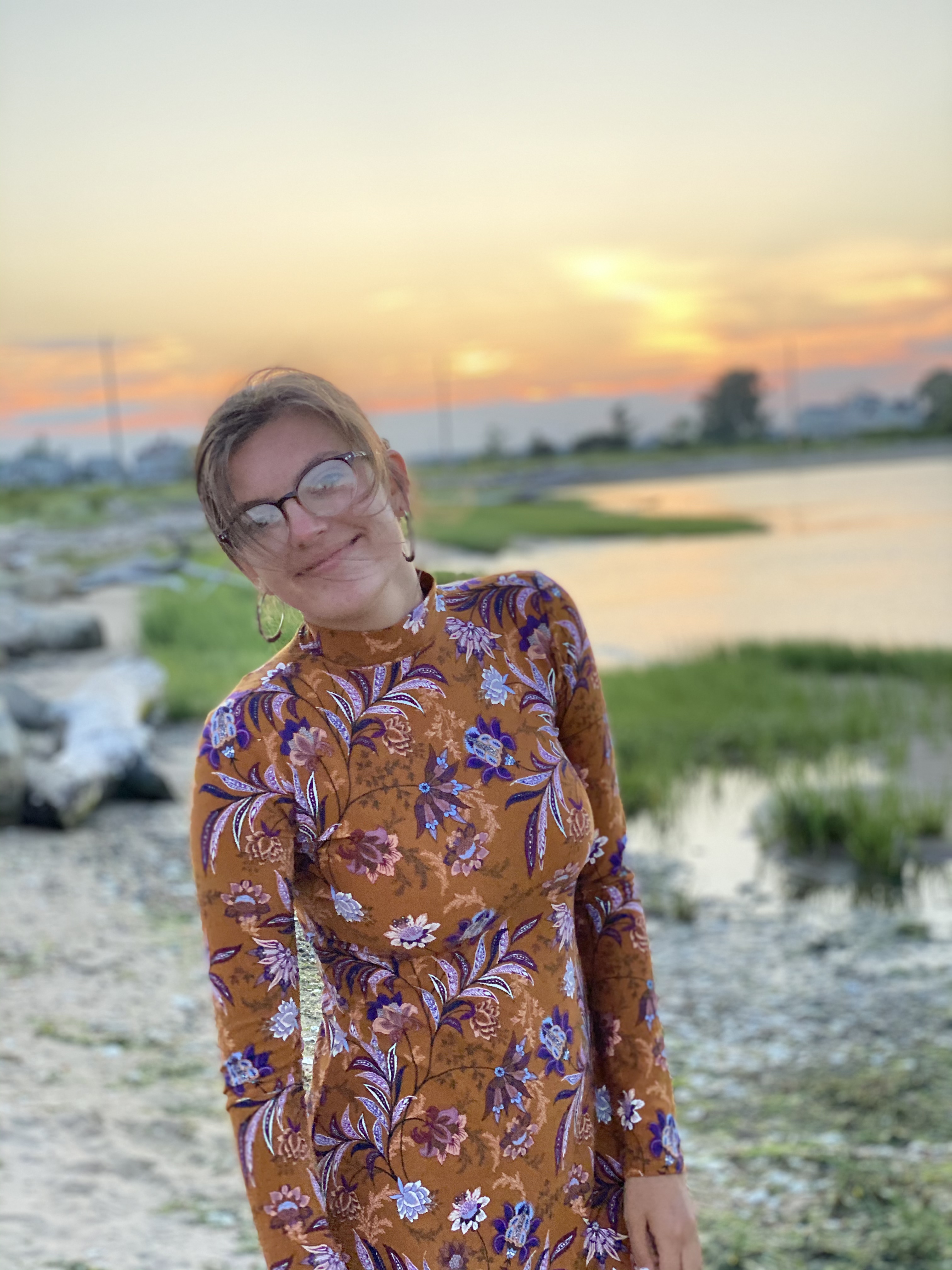 Paleena, wearing glasses and a brown and purple floral dress with a sunset behind her. 
