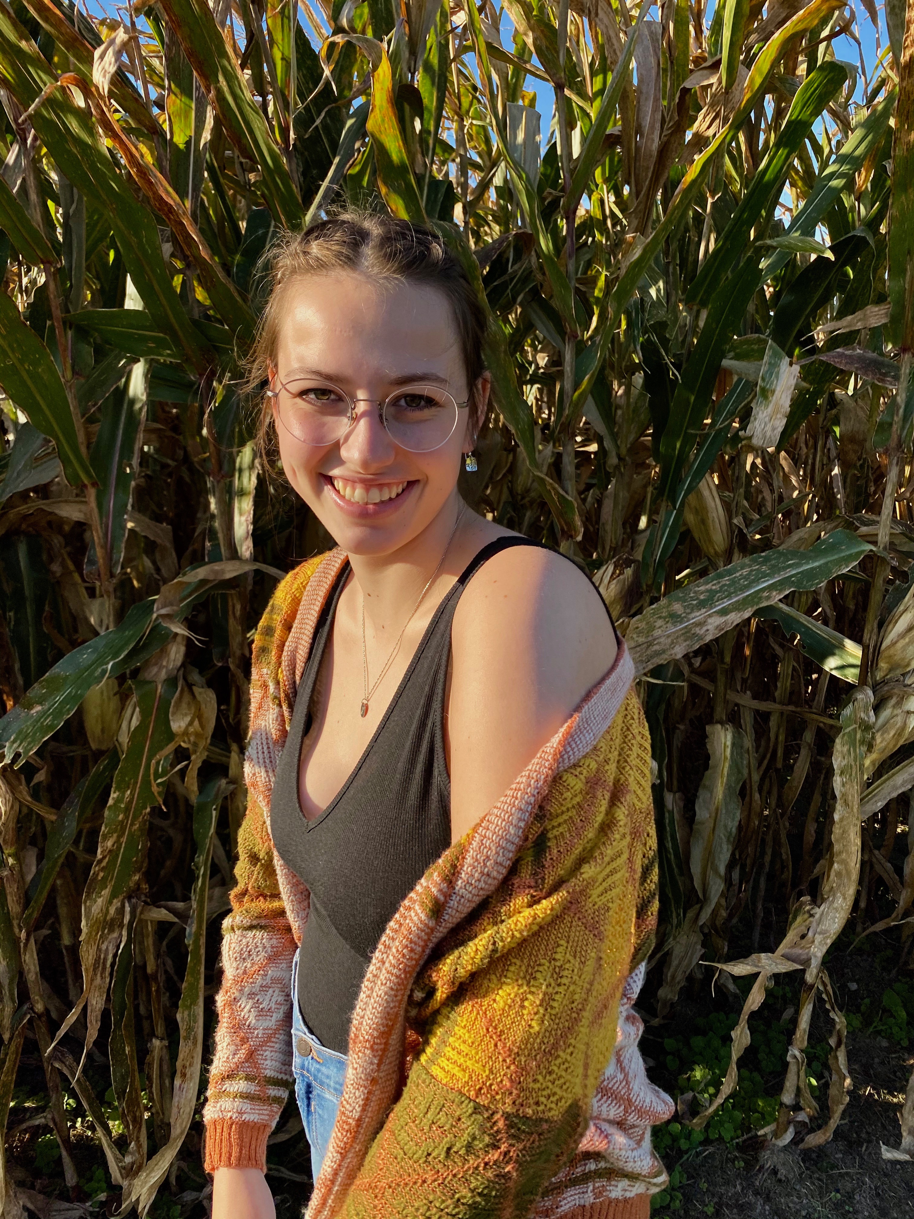 Sylvie, wearing a black shirt and multicolored cardigan, smiling with a field of corn behind her. 